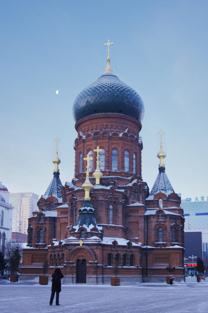 Snow-covered Saint Sophia Cathedral in Harbin photographed at dusk, with a lone photographer standing in the foreground under a pale winter sky.