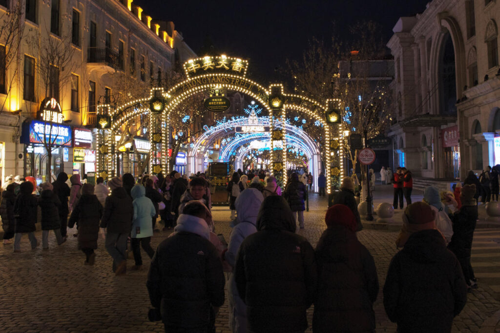 Crowds walk beneath illuminated arches at the entrance of Zhongyang Street in Harbin on a winter night.