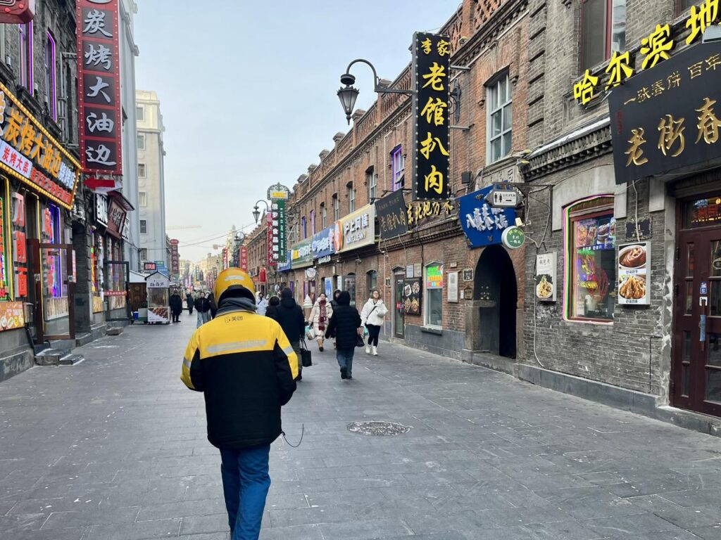 A food delivery rider walks through a street in Daowai District, Harbin, lined with Chinese Baroque buildings and restaurant signs.