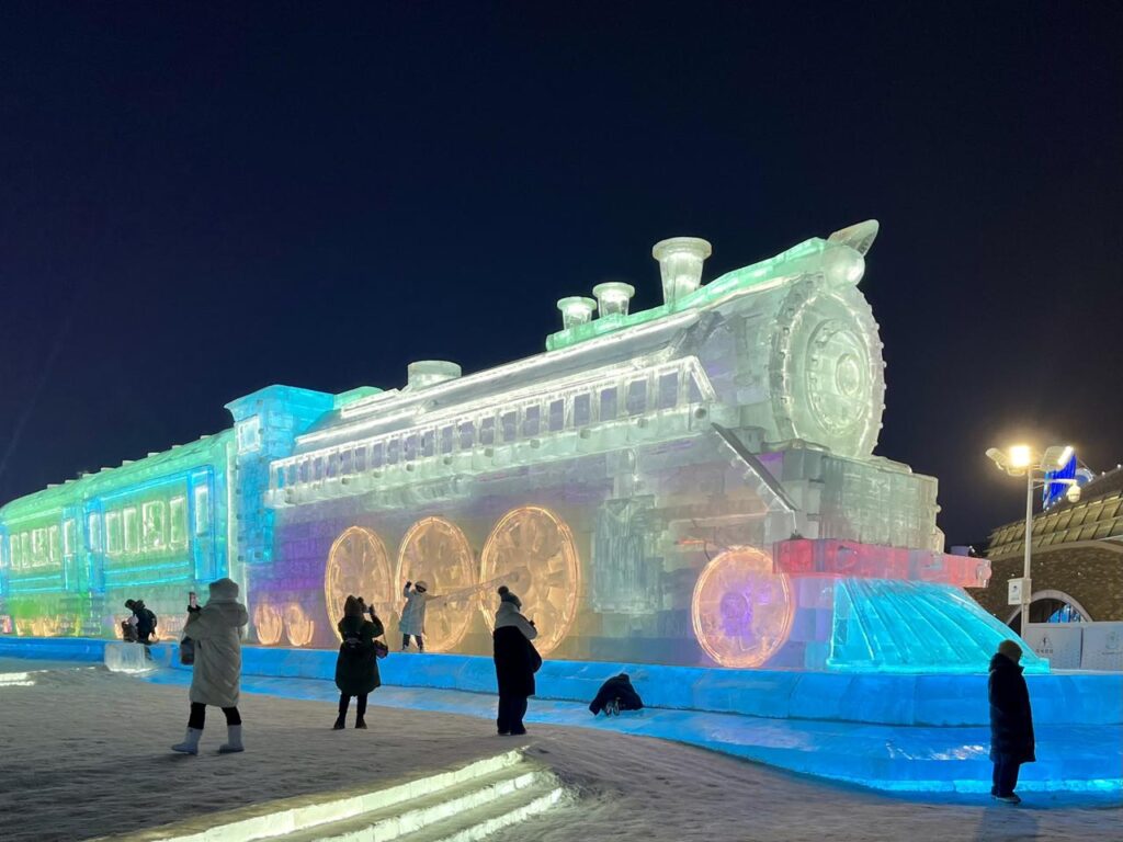 Visitors walk past a large illuminated ice sculpture shaped like a steam locomotive at Ice and Snow World in Harbin at night.