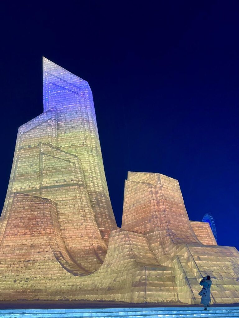 A towering ice sculpture illuminated in warm and cool colors at Harbin Ice and Snow World, with a single visitor standing at its base for scale.