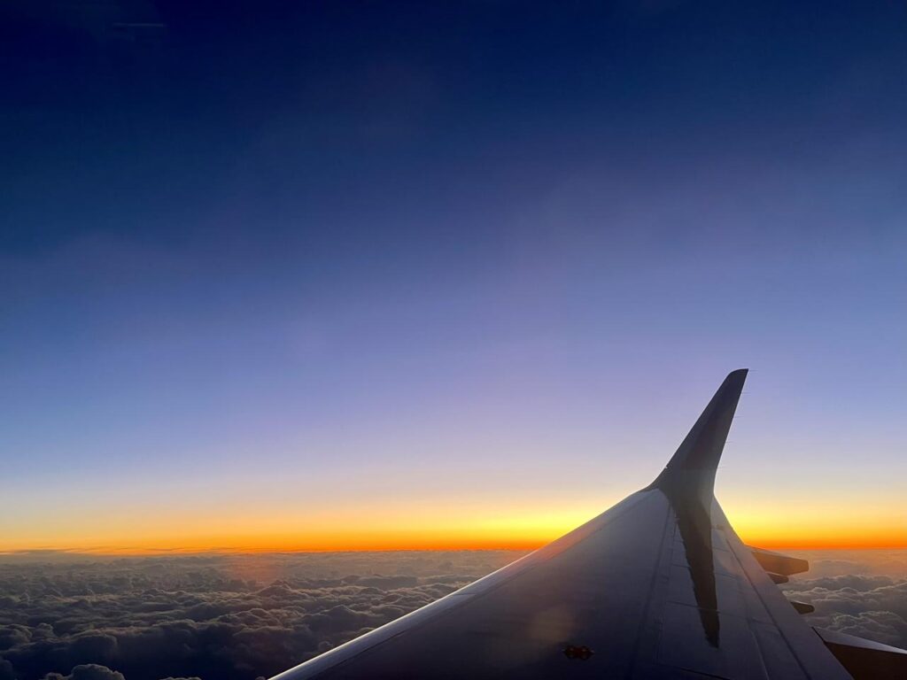 Sunset viewed from an airplane window, with the aircraft wing silhouetted against glowing clouds and a fading blue sky.